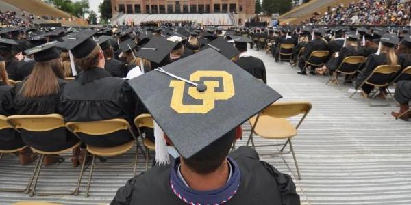 A view of the stage from the back of the student section at the spring commencement ceremony. In the foreground, a student is wearing a graduation cap with the CU logo on the top.